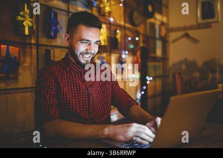 Theme training and computers. A young man with a beard and in a shirt uses a laptop, prints on the keyboard in a coffee shop at a wooden table in the Stock Photo