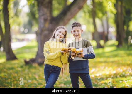 Happy Caucasian siblings enjoying in autumn day in park, sunny weather. Kids having fun holding fallen yellow leaves in nature. Funny twins in autumn Stock Photo