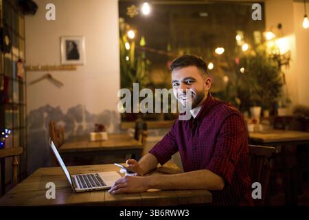 A young handsome Caucasian man with beard and toothy smile in red checkered shirt is working behind laptop sitting at wooden table. Uses holds mobile Stock Photo