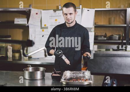 Theme cooking. A young Caucasian man in black uniform and latex gloves at a restaurant in the kitchen. Cooking mincers into a berry with a syringe. A Stock Photo