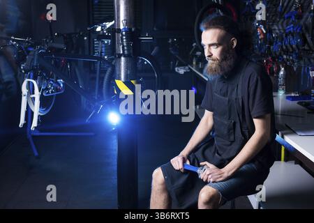 Portrait of small business owner of young man with beard. Guy bicycle mechanic workshop worker sitting with tool in his hand in a working black clothe Stock Photo