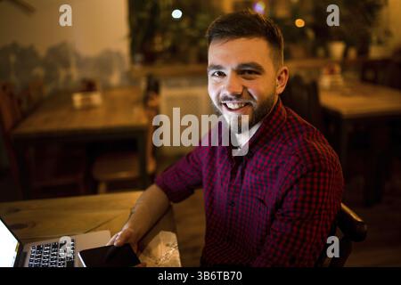 A young handsome Caucasian man with beard and toothy smile in red checkered shirt is working behind laptop sitting at wooden table. Uses holds mobile Stock Photo