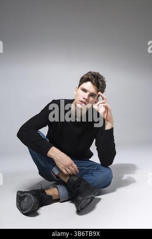 Stylish guy posing in studio sitting on the floor in denim pants and black sweatshirt. Male student portrait full on white background. Studio shot Han Stock Photo
