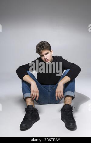 Stylish handsome man posing sitting on floor. Handsome Guy. cool fashion male model sitting on grey background and looking at the camera. Student in a Stock Photo