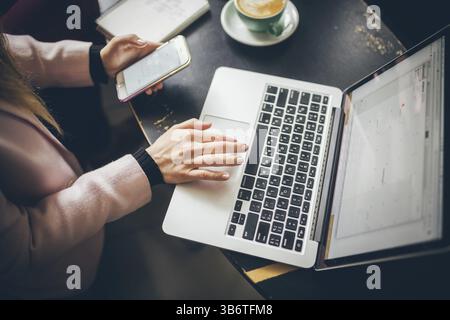 Close-up top view of Caucasian woman's hands Casually dressed student, blogger, writer man working on a laptop holding a phone in his hand, inside the Stock Photo