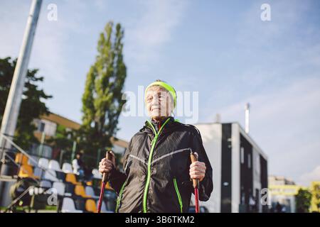 Senior woman walking with walking poles in stadium on a red rubber cover. Elderly woman 88 years old doing Nordic walking exercises at the city stadiu Stock Photo