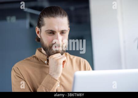 A bearded man with a ponytail looks down at a laptop, pondering a thought while working in an office setting. Stock Photo
