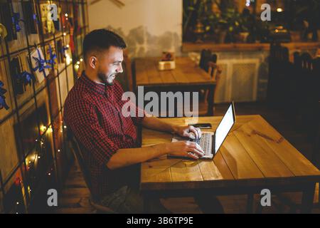 A young handsome Caucasian man with beard and toothy smile in a red checkered shirt is working behind a grey laptop sitting at a wooden table. Hands o Stock Photo