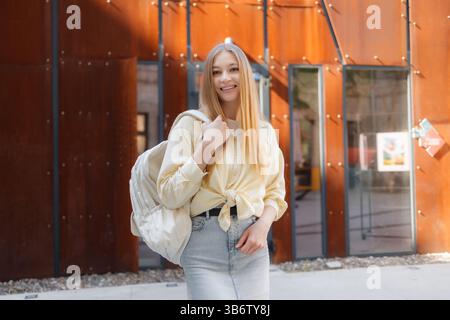 Young woman smiles while standing in front of modern architecture during the day with a backpack Stock Photo