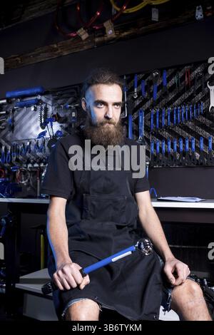 Portrait of small business owner of young man with beard. Guy bicycle mechanic workshop worker sitting with tool in his hand in a working black clothe Stock Photo