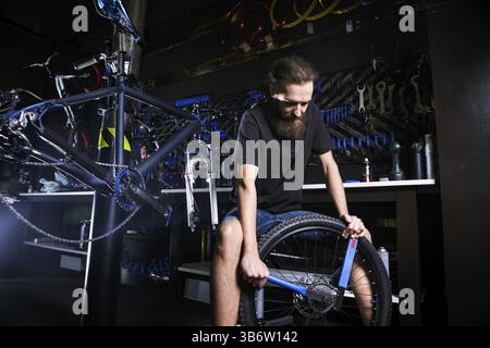 Young stylish bearded, with long hair Caucasian male mechanic worker at a bicycle workshop uses a tool for repairing Cassettes on the rear wheel of a Stock Photo