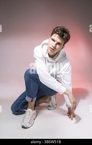 Handsome young stylish man sitting on the floor in studio. Teenager model posing while sitting on a pink background in casual clothes. The guy in jean Stock Photo