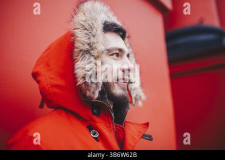 Portrait of a young handsome guy, a student with a beard in a red winter jacket and a hood with fur on the background of the red wall of the building Stock Photo