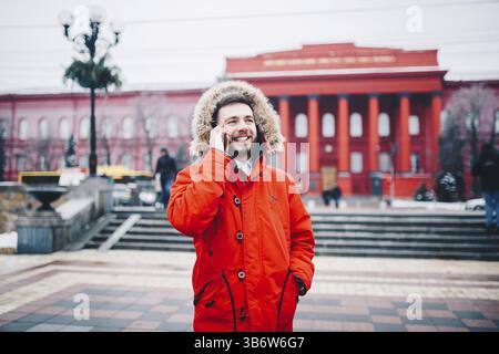 Young guy with beard and red jacket in hood a student uses mobile phone, holds in his hand near the head, talking on the phone with smile on the backg Stock Photo
