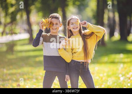 Happy Caucasian siblings enjoying in autumn day in park, sunny weather. Kids having fun holding fallen yellow leaves in nature. Funny twins in autumn Stock Photo