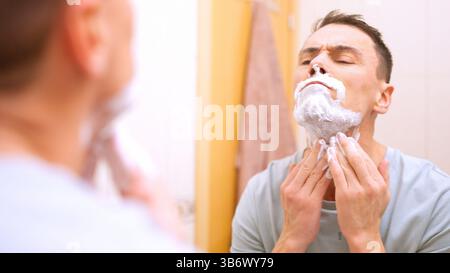 Man applying shaving cream on face in bathroom mirror Stock Photo