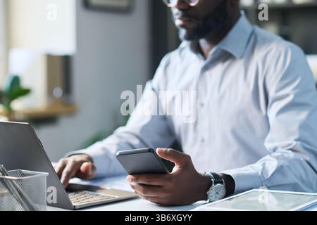 Black Male Company Director Using Smartphone and Laptop in Office Stock Photo