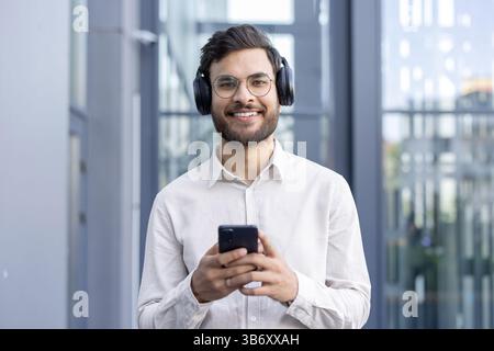 A smiling man wearing headphones and glasses holds his smartphone, possibly listening to music in an office setting. Stock Photo