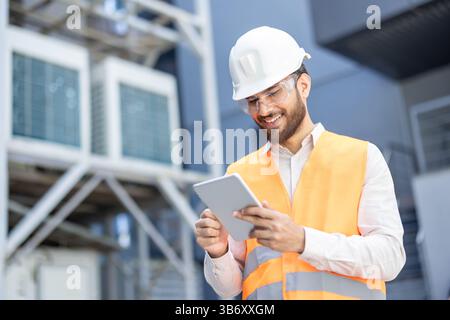 A smiling engineer in a white helmet and safety vest uses a tablet in a industrial setting, possibly for equipment inspections. Stock Photo