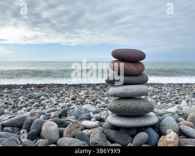Balanced stack of smooth sea stones on a pebble beach with ocean waves and cloudy sky in the background. No people. Stock Photo