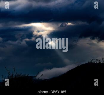 A dramatic sky filled with dark clouds, illuminated by a single beam of sunlight breaking through. The landscape features a silhouette of a mountain a Stock Photo