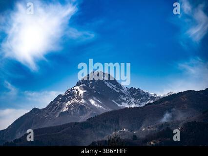 A majestic mountain peak covered with snow, under a bright blue sky with wispy clouds. The lower slopes are forested, and a few small houses can be se Stock Photo