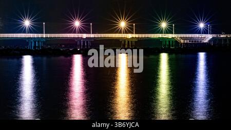 A nighttime view of a pier illuminated by colorful lights reflecting on the water's surface. The scene captures a serene atmosphere with vibrant starb Stock Photo
