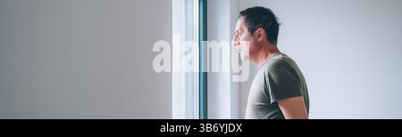 Serious caucasian male looking through window and thinking in morning, selective focus Stock Photo