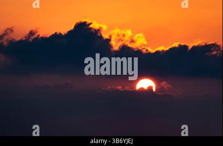 A stunning sunset with the sun partially hidden behind dark clouds, casting a warm orange glow in the sky. The scene evokes tranquility and the beauty Stock Photo
