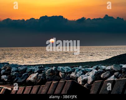 A serene sunset over the ocean, with the sun partially hidden behind dark clouds. The foreground features a rocky shoreline and wooden structures, cre Stock Photo