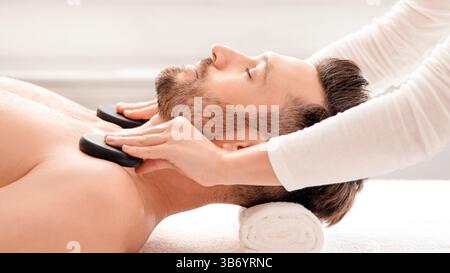 Spa female therapist placing black stones on relaxed man shoulders Stock Photo