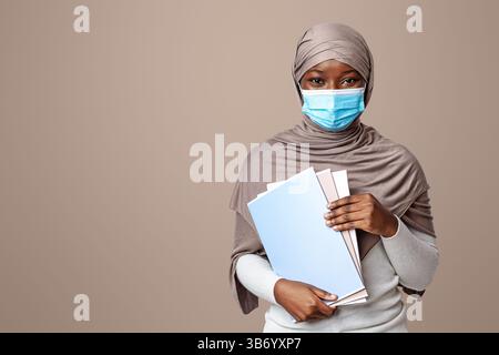 Young black muslim female student in medical mask posing with workbooks Stock Photo