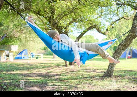 Young hispanic man resting at midday under the shade of trees in a hammock hanging outdoors at a campsite. Stock Photo
