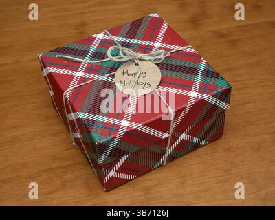 A charming red plaid gift box, wrapped with rustic twine and a Happy Holidays tag, rests on a wooden surface, radiating warmth and festive cheer. Stock Photo