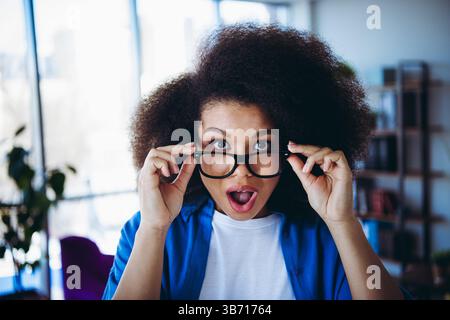 Young woman in a modern office environment looking surprised while adjusting her glasses Stock Photo