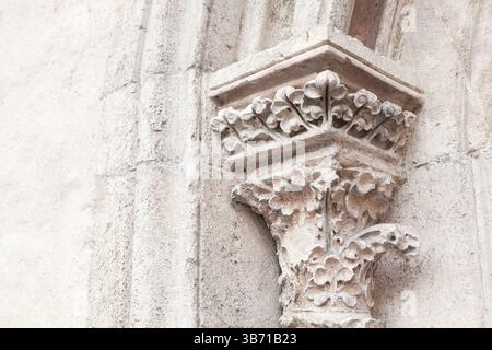 Intricately carved stone capital adorns top of column on  historic building, featuring floral and leaf motifs. Weathered texture of stone contrasts Stock Photo