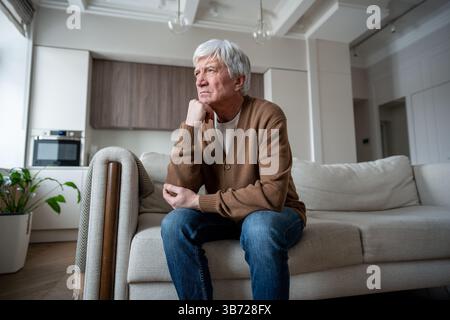 Thoughtful struggling elderly man rests fist against cheek, looking out window alone on sofa Stock Photo
