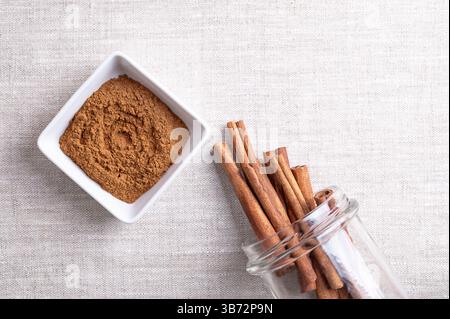Cassia cinnamon powder in a white bowl, with cinnamon sticks sticking out of an open screw-top jar, on linen fabric, with empty space for text. Stock Photo