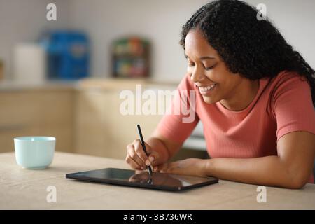 Happy black woman drawing with stylus pen in a tablet in the kitchen at home Stock Photo