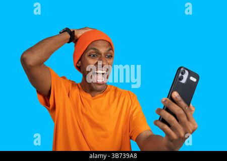 Young man expressing excitement while using smartphone against a vibrant blue backdrop Stock Photo