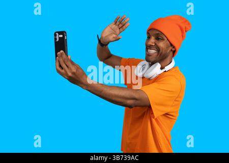Young man wearing headphones making a video call with his smartphone and waving on a blue background Stock Photo