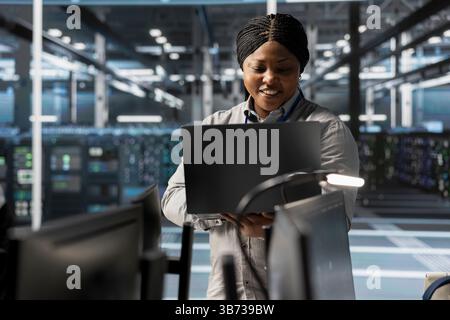 Smiling engineer using laptop in data center to oversee hardware delivering computing power for AI training models. Happy worker supervising rackmount servers storing complex machine learning datasets Stock Photo