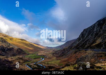 Meandering Afon Ogwen threading through heather-clad glacial valley beneath a dynamic Snowdonia sky Stock Photo