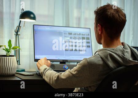 Man in gray hoodie working at computer desk with dual monitors and lamp in background. Freelancer working remotely at home office. Concept of remote w Stock Photo