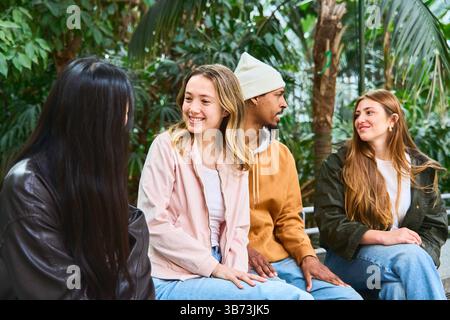 Four university students enjoying a conversation together, sitting and relaxing in a botanical garden full of lush plants Stock Photo