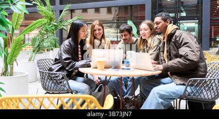 Group of diverse university students working together on laptops, enjoying coffee and collaborating on a project in a modern cafe courtyard Stock Photo