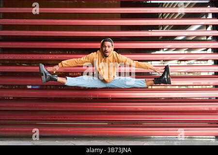 Young man wearing casual clothes jumping doing the splits while making funny face in front of a red metallic roll up door Stock Photo