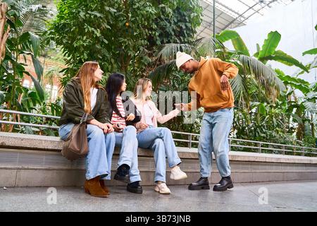 Young man greeting three female friends sitting on a bench in a botanical garden, talking and having fun Stock Photo