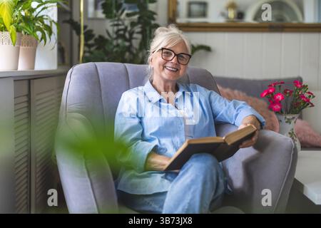 Mature woman reading a book in a comfortable armchair at home Stock Photo