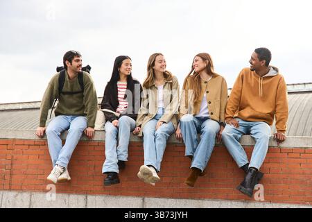 Five multiethnic young students are enjoying each other's company, sitting on a low brick wall, engaging in lively conversation and sharing laughter Stock Photo